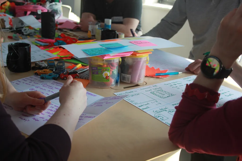 Close-up of hands writing on paper worksheets surrounded by craft supplies and sticky notes on a table